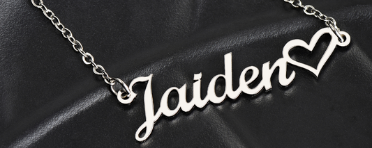 A close-up of a personalized jewelry piece featuring a silver heart name necklace with elegant script and a heart detail at the end, displayed on a black leather surface.