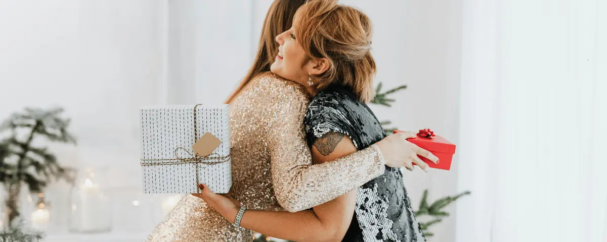 Two women hugging beside wrapped presents, one holding a red personalized gift box